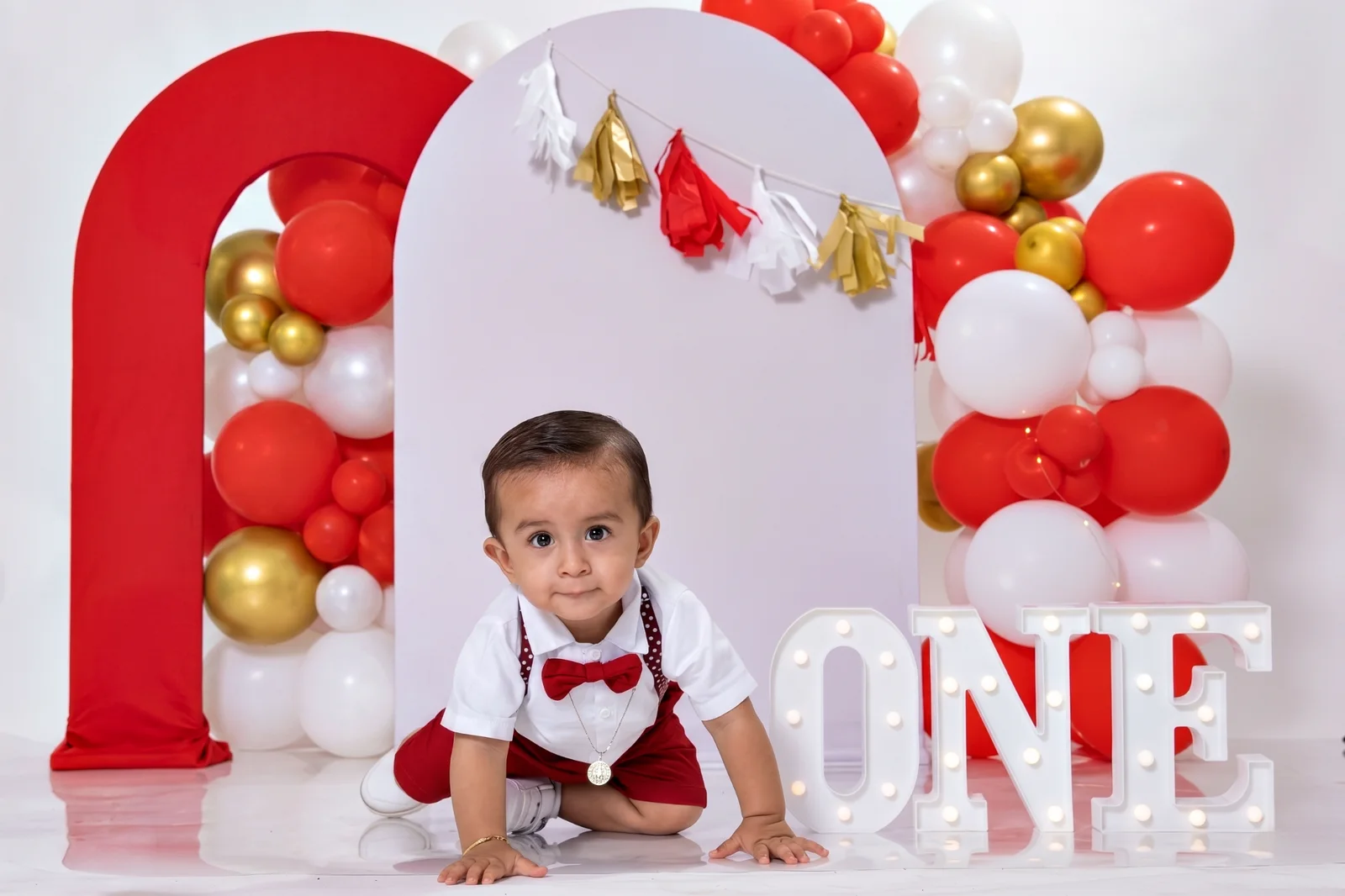 Bebé gateando frente a globos rojos, blancos y dorados en su sesión de primer cumpleaños.