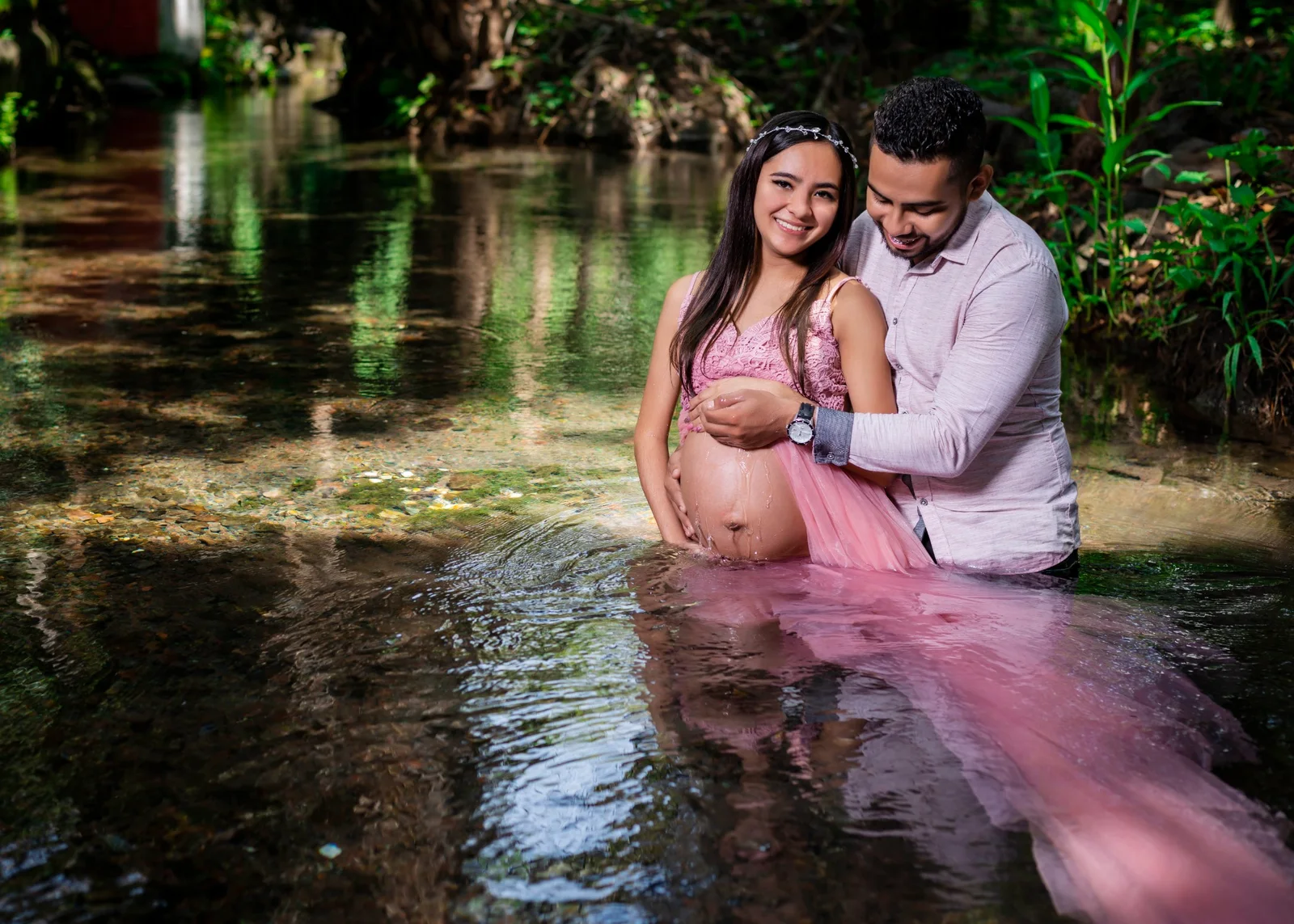 Pareja en sesión de maternidad dentro del agua con vestido rosa.
