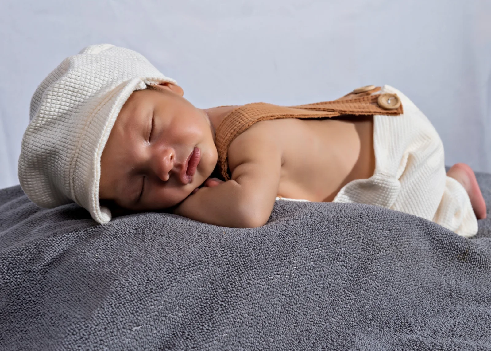Bebé dormido de perfil con gorro blanco y conjunto en tonos crema y café.