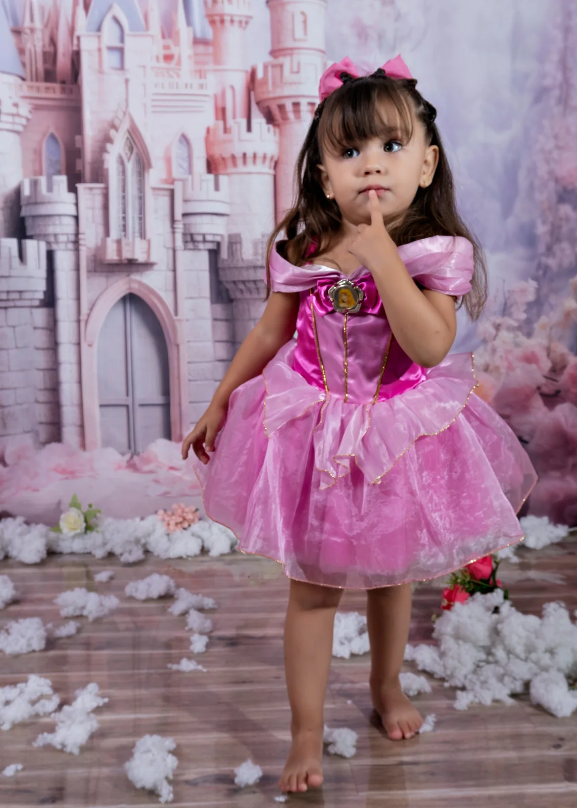 Niña con vestido de princesa rosa y un dedo junto a los labios frente a un castillo.